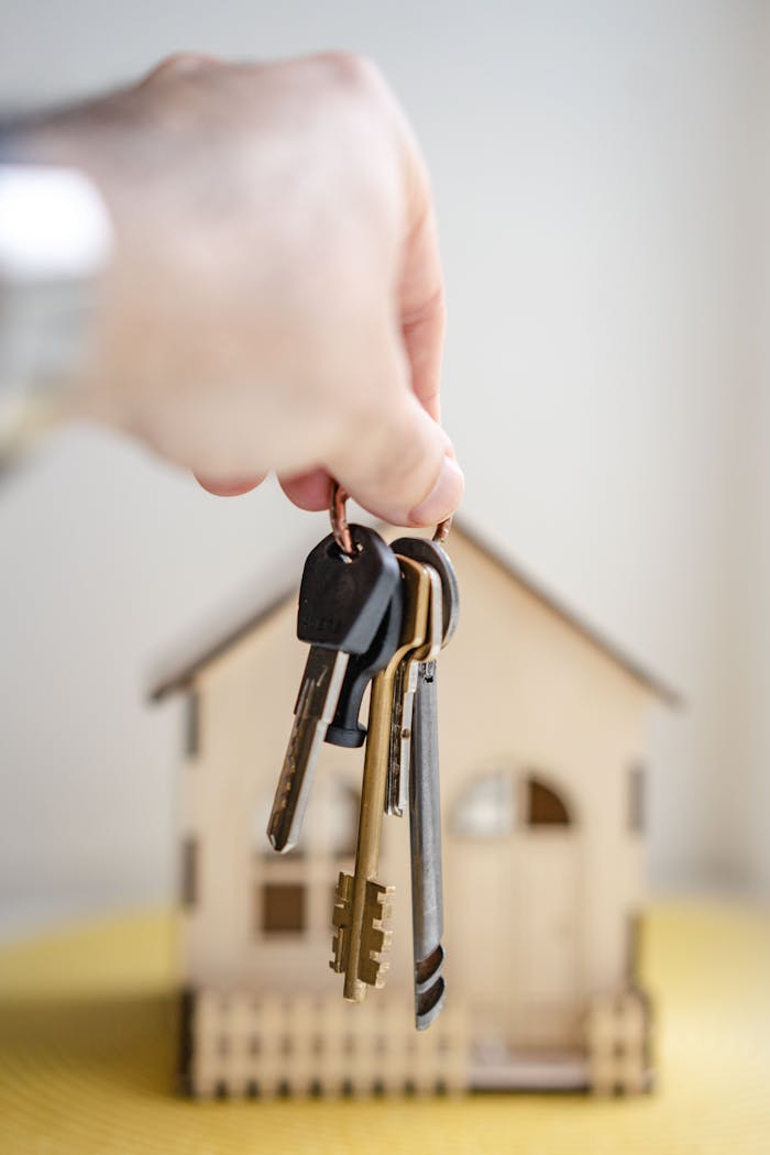 gallery-2 Close-up of a hand holding keys with a miniature wooden house in the background, symbolizing real estate investment.